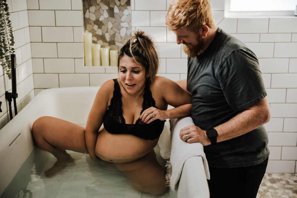 woman in tub feeling for baby's head as baby exits while husband watches her